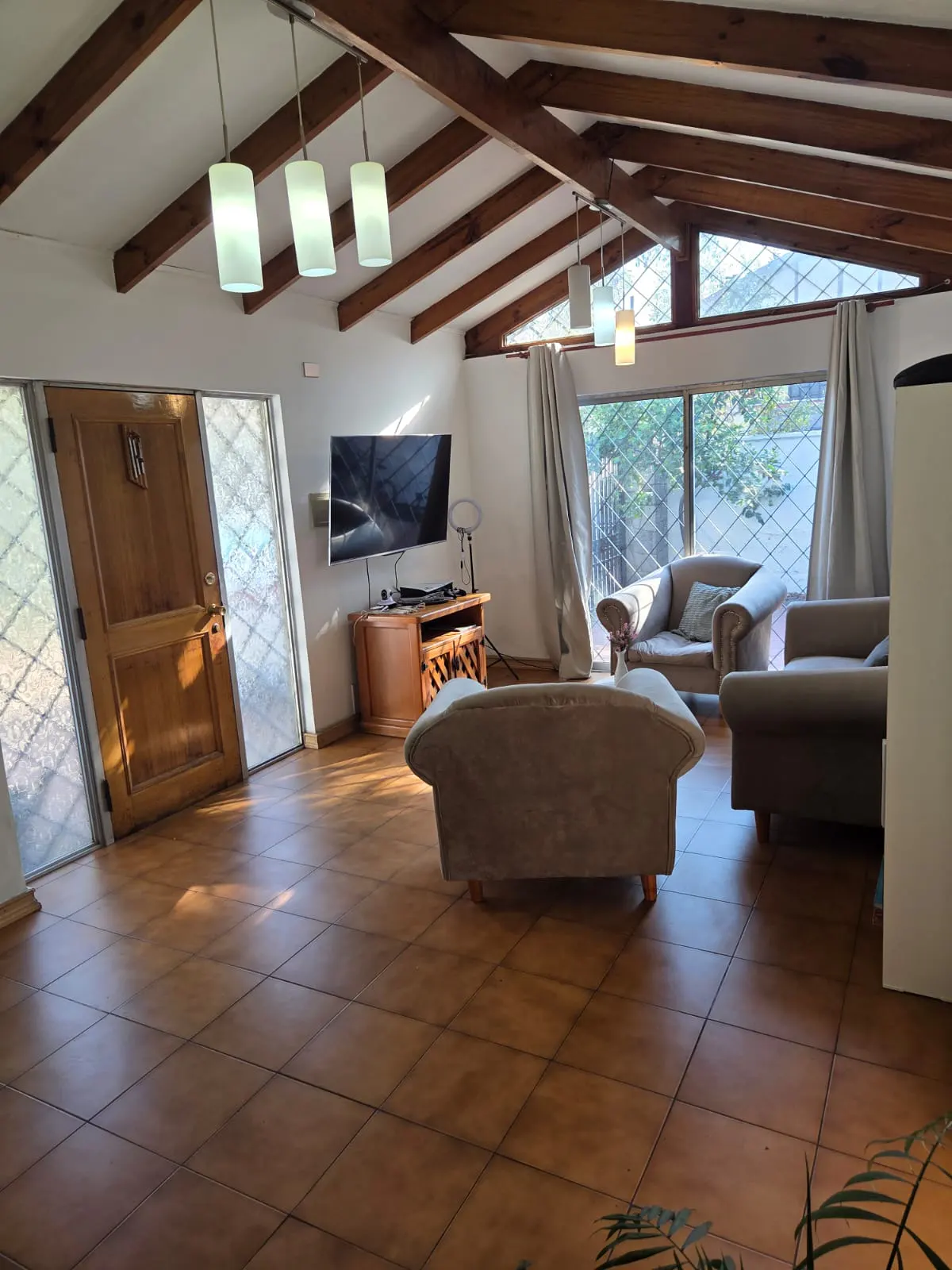 Living room with vaulted wood-beam ceiling, tile floors, beige sofas, a wall-mounted TV, and large windows with curtains.