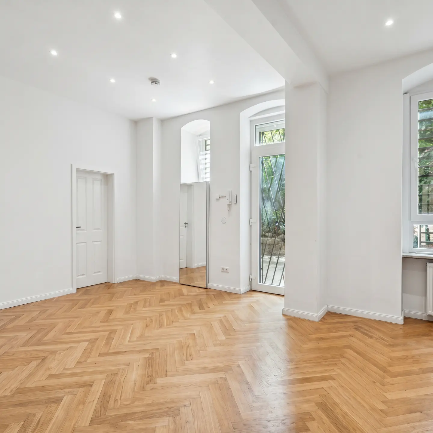 Bright, empty room with white walls, herringbone wood floor, door, mirror, and windows with diamond pattern.