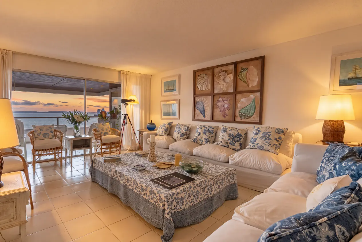 Living room with white sofa, blue patterned pillows, and a coffee table with a blue patterned tablecloth. Balcony with ocean view.