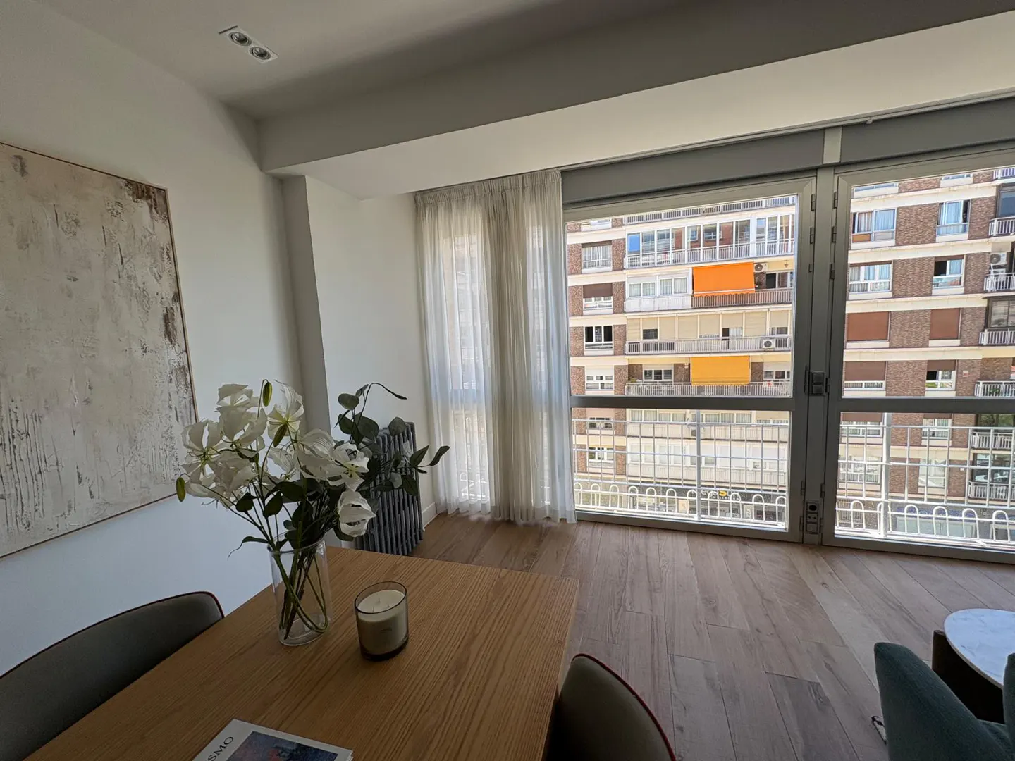 Modern dining area with a wooden table, white flowers, and large windows overlooking a city apartment building.
