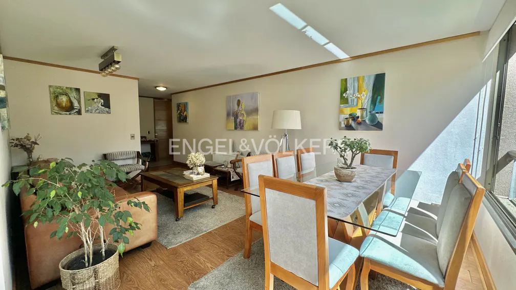 Bright living room with wood floors, a glass dining table with six chairs, and a brown leather couch. Paintings decorate the walls.
