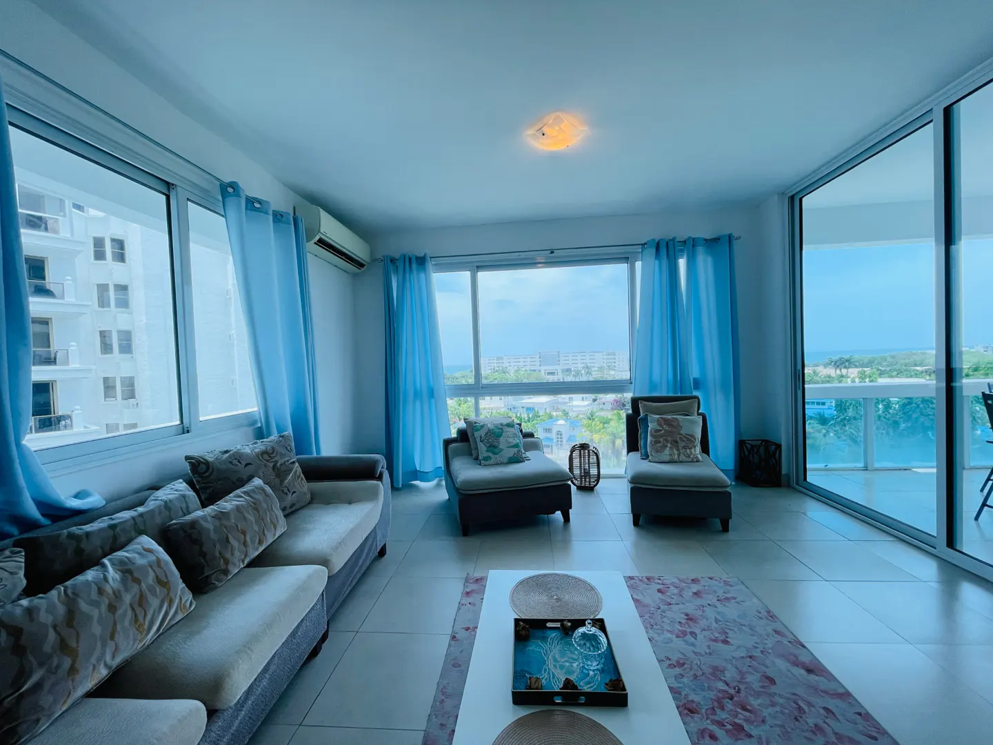 Bright living room with a sofa, two chaise lounges, and a white coffee table. Blue curtains frame large windows with city and ocean views.
