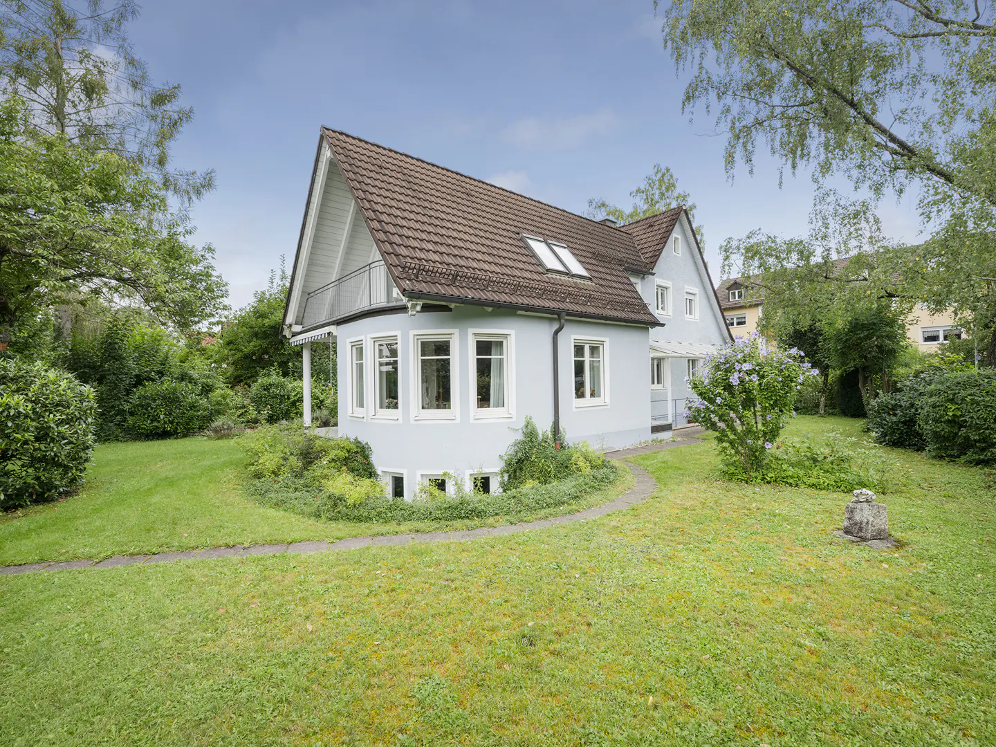 Light blue house with a brown roof, surrounded by a green lawn and trees under a blue sky.