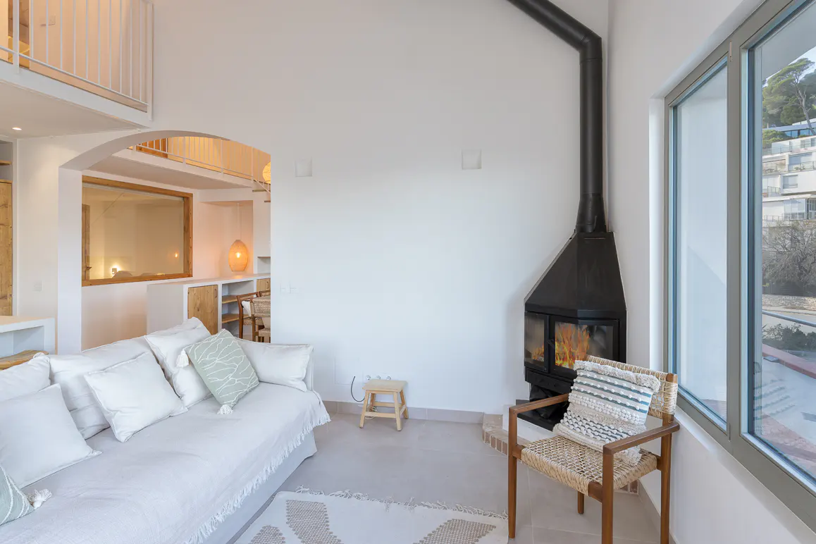 Bright living room with white walls, a black fireplace, and a large window with an outdoor view. A white sofa and woven chair add cozy seating.