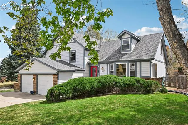 Two-story house with gray roof, white siding, red door, and two-car garage, surrounded by green lawn and bushes.
