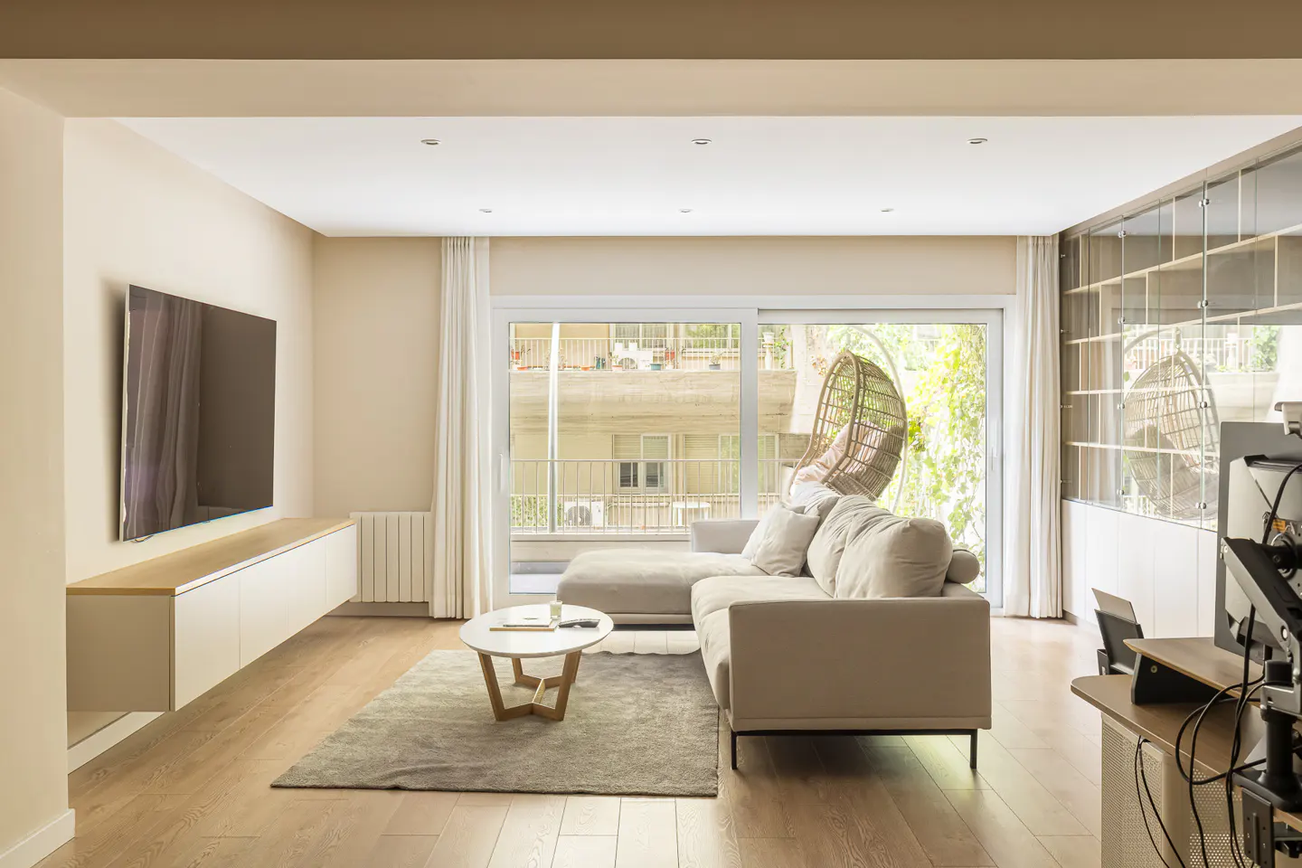 Bright living room with hardwood floors, a beige sectional sofa, and a large sliding glass door to a balcony. A TV is mounted above a white cabinet.