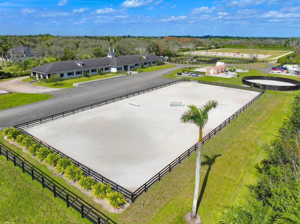 Aerial view of a horse farm with a large white riding arena, black fences, and a long white barn under a blue sky.