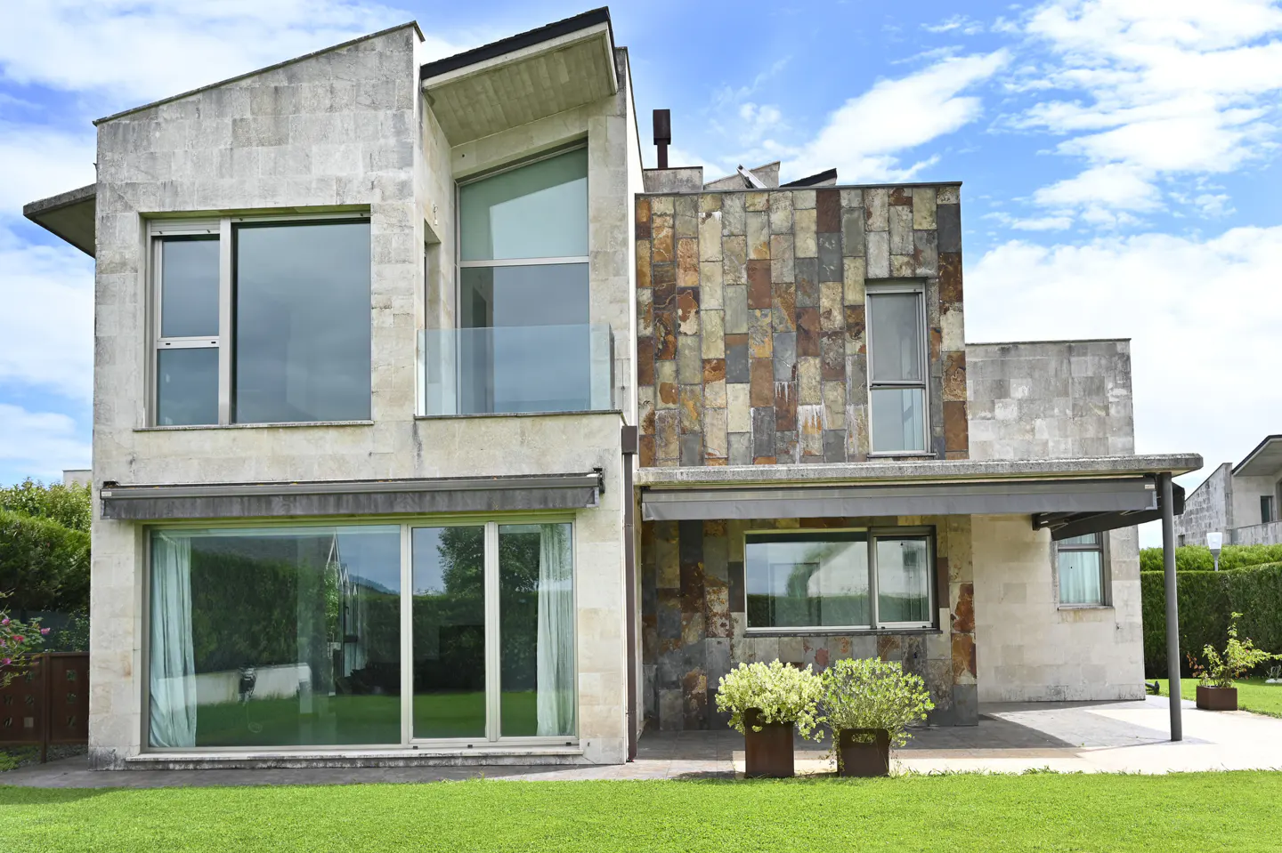 Modern two-story house with stone and tile exterior, large windows, and green lawn under a blue sky.