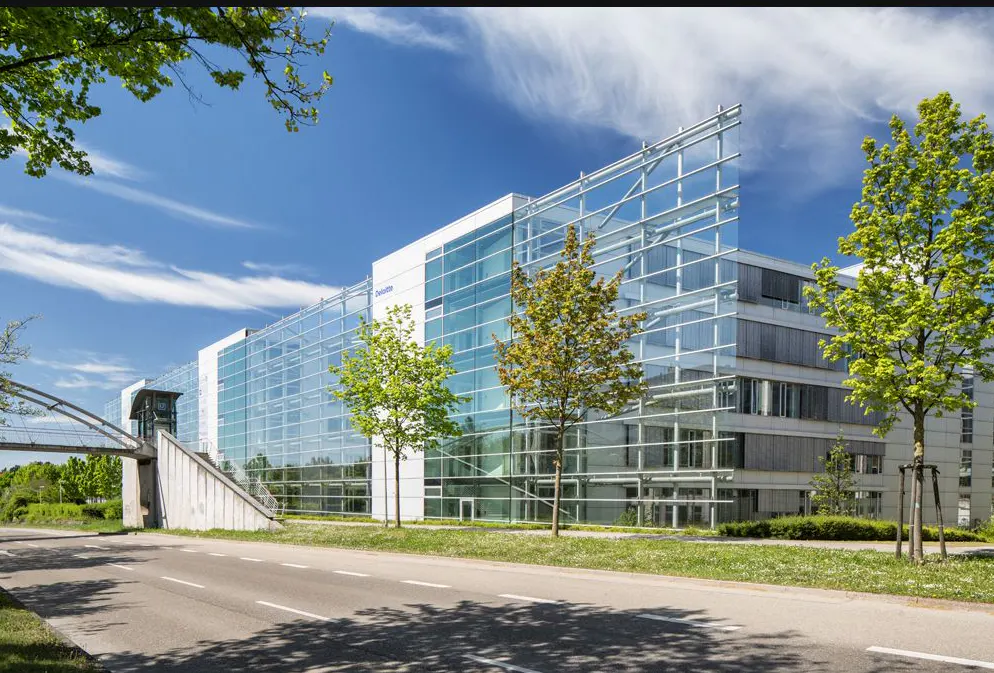 Modern glass and white office building with a skybridge, trees, and a road in front.