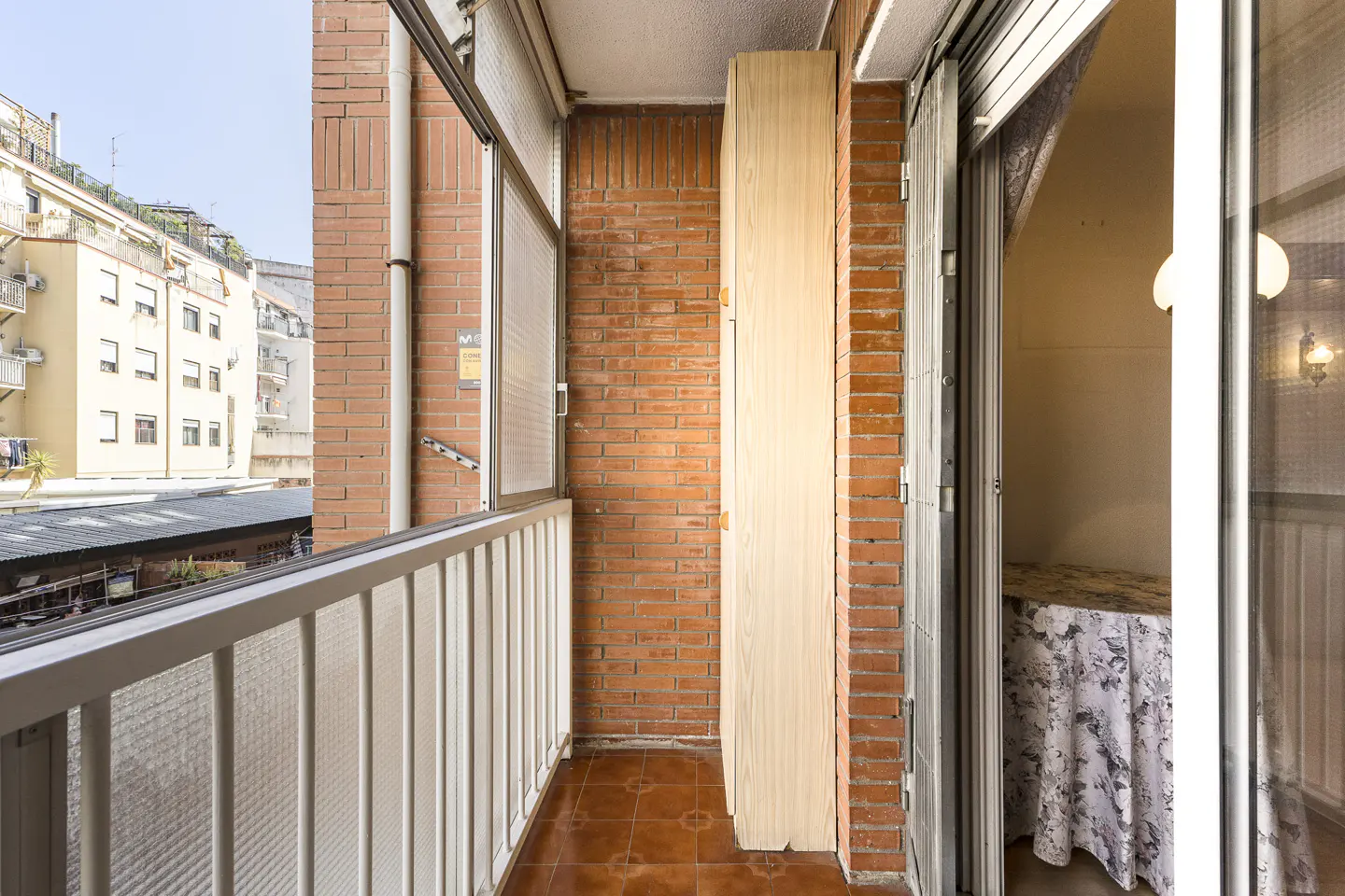 A balcony with a white railing, brick walls, and brown tile floor. A tall, light-colored cabinet stands against the brick. Sliding glass doors open to a room with a table.