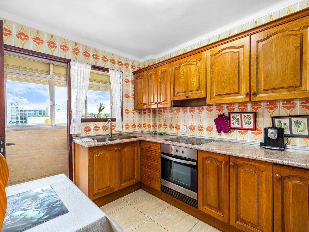 A kitchen with wood cabinets, orange patterned wallpaper, and a stainless steel oven. A window with white curtains overlooks a balcony.