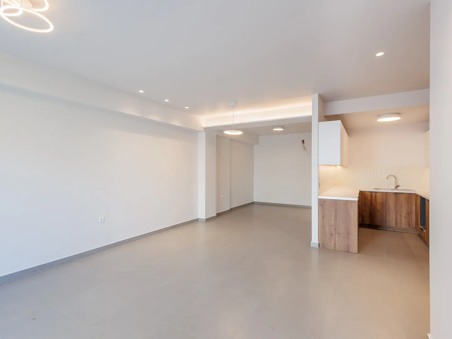 Modern open-plan living area with white walls, light grey floors, and a kitchen featuring wood cabinets and white counters.