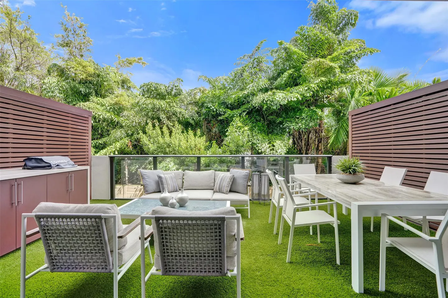 Outdoor patio with white furniture, a dining table, and a sofa on green turf. Lush green trees and a blue sky are in the background.