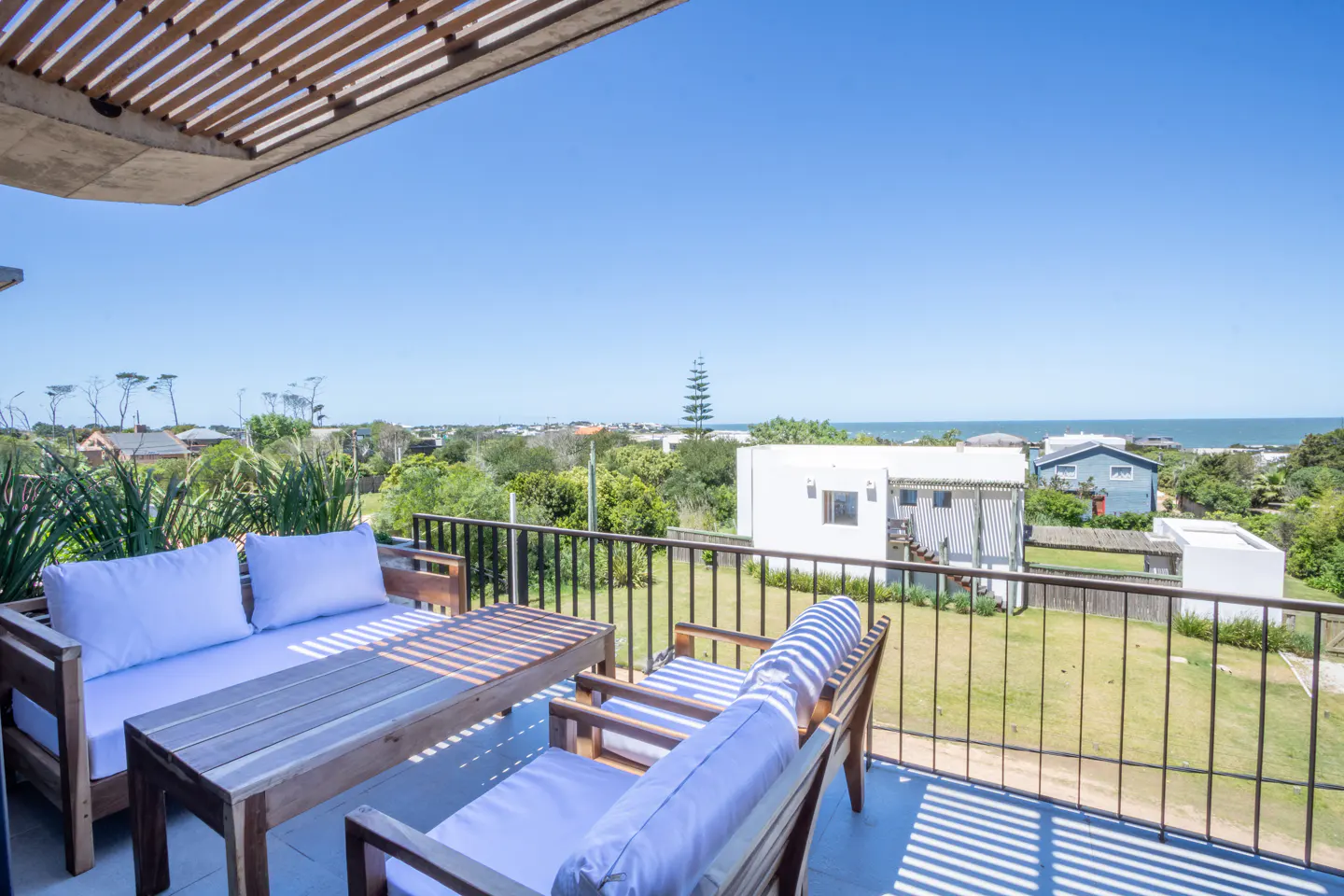 Outdoor patio with wood furniture and blue cushions overlooks a green lawn, houses, trees, and the ocean under a clear blue sky.