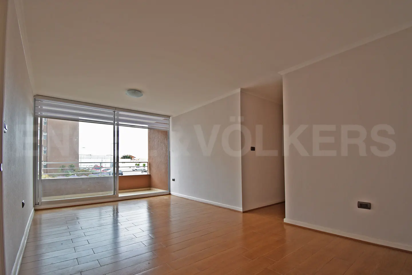 Empty room with wood floors, white walls, and a sliding glass door to a balcony. "VÖLKERS" is printed on the wall.