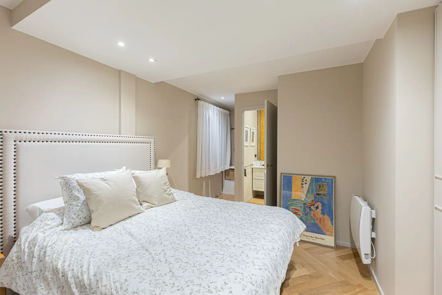 Bedroom with a white upholstered bed, patterned duvet, and herringbone wood floor. A bathroom is visible through an open door.