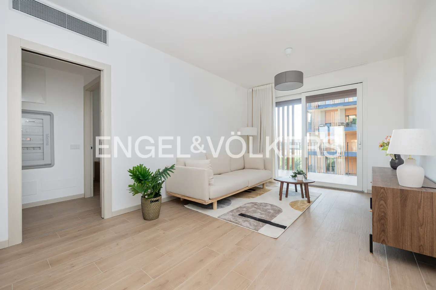 Bright living room with wood floors, white walls, and a beige sofa. A sliding glass door leads to a balcony. A potted plant sits near the sofa.