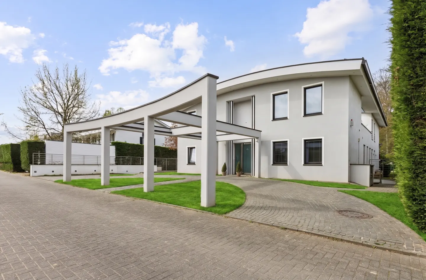 Modern gray house with a curved roofline and carport. A paved driveway leads to the entrance. Green grass and trees surround the property.