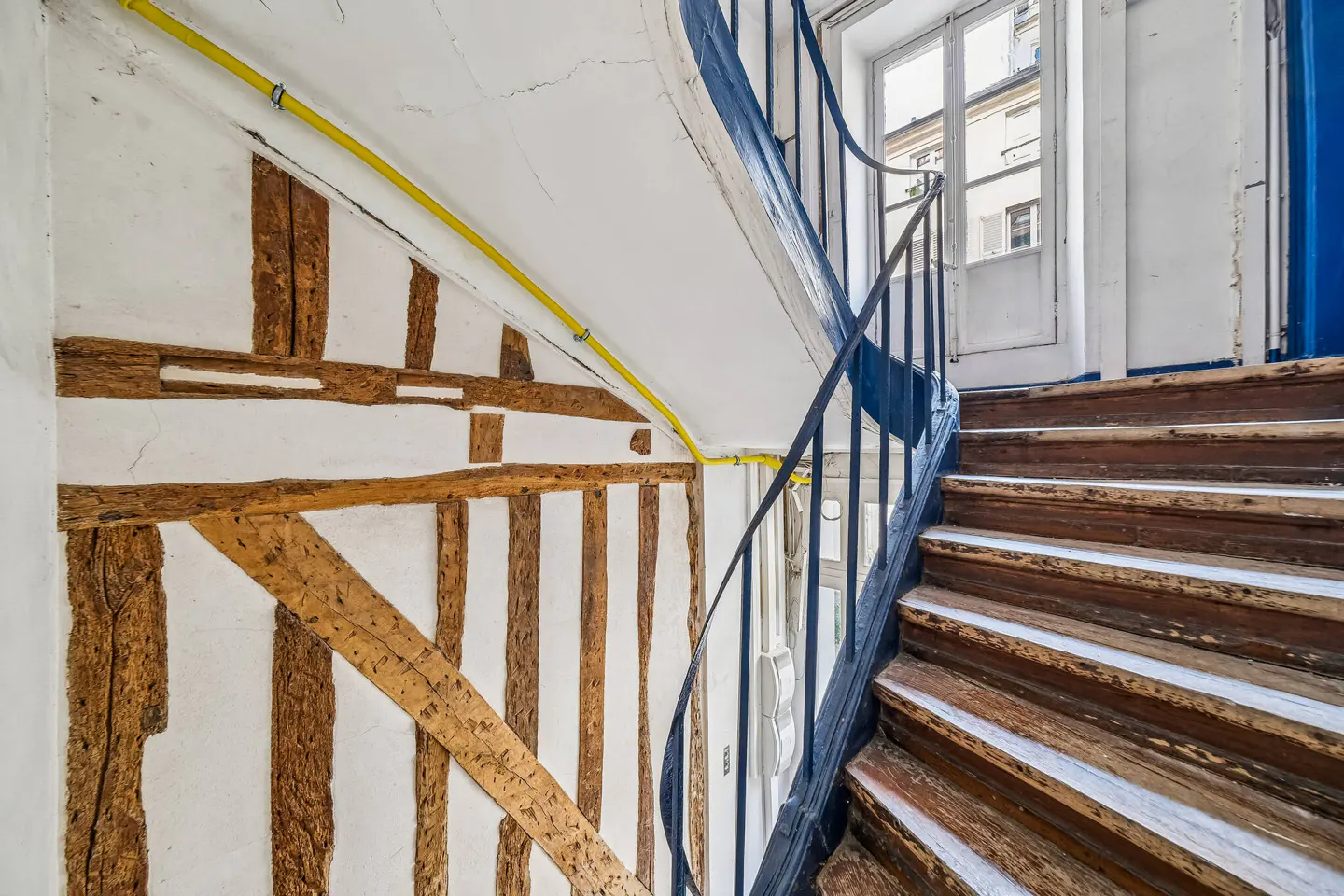 Interior view of a wooden staircase with a blue railing, next to a white wall with exposed timber framing and a yellow pipe.