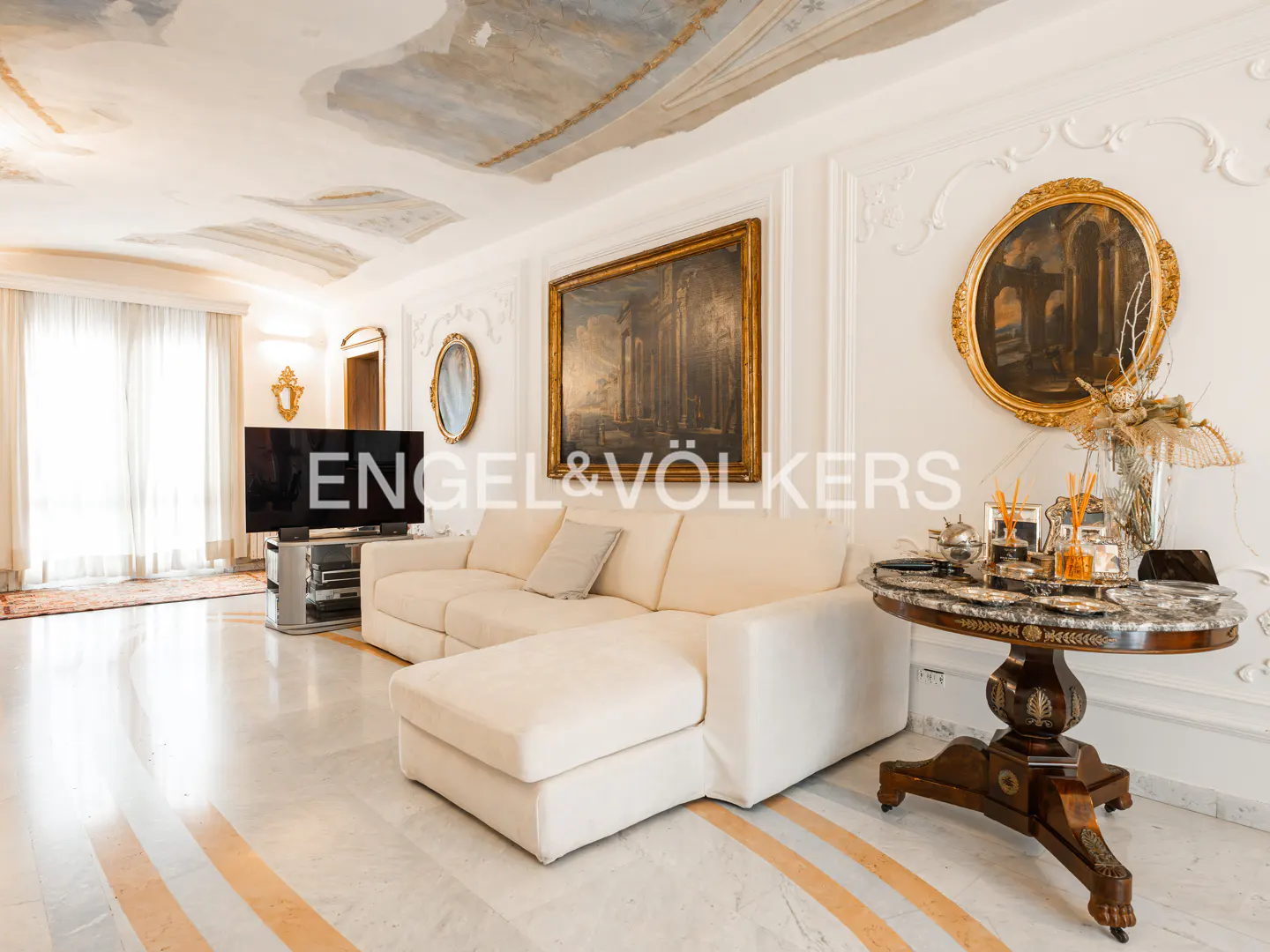 A bright living room with a white sectional sofa, marble floors, and ornate gold-framed art.