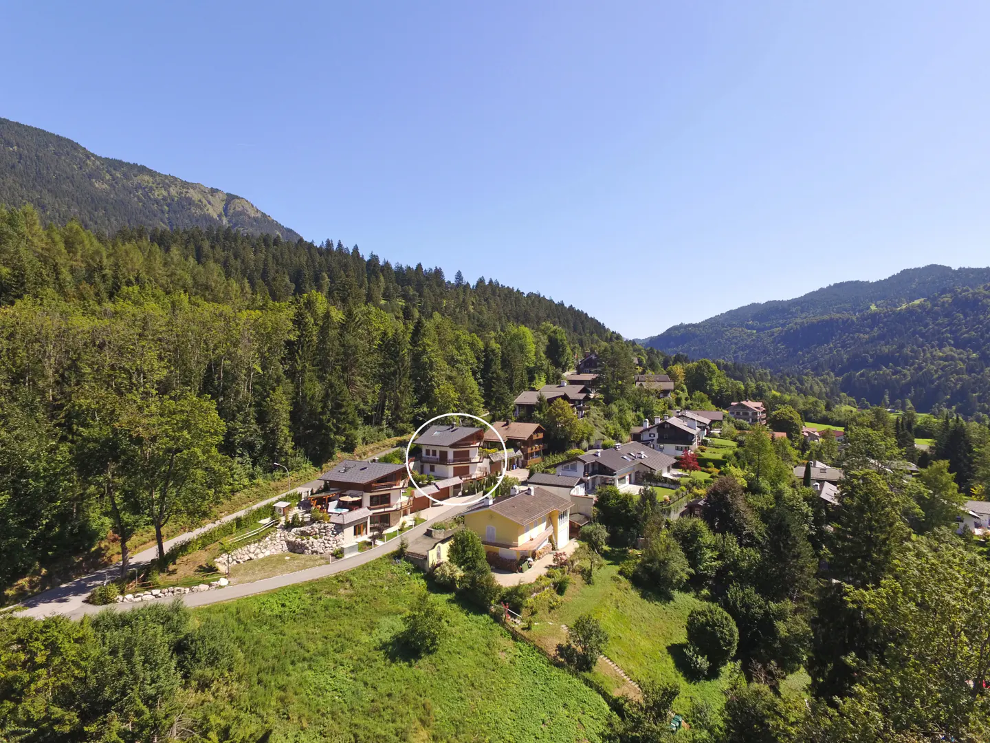 Aerial view of a multi-story house with a brown roof, surrounded by green trees and mountains under a blue sky.