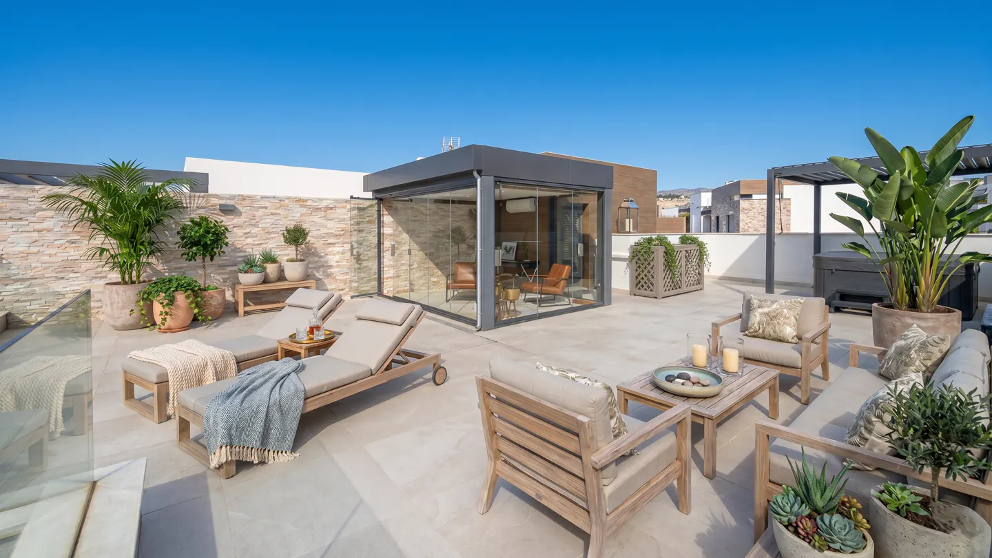 Rooftop patio with lounge chairs, sofa, and enclosed sunroom. Plants in pots add greenery. Blue sky above.