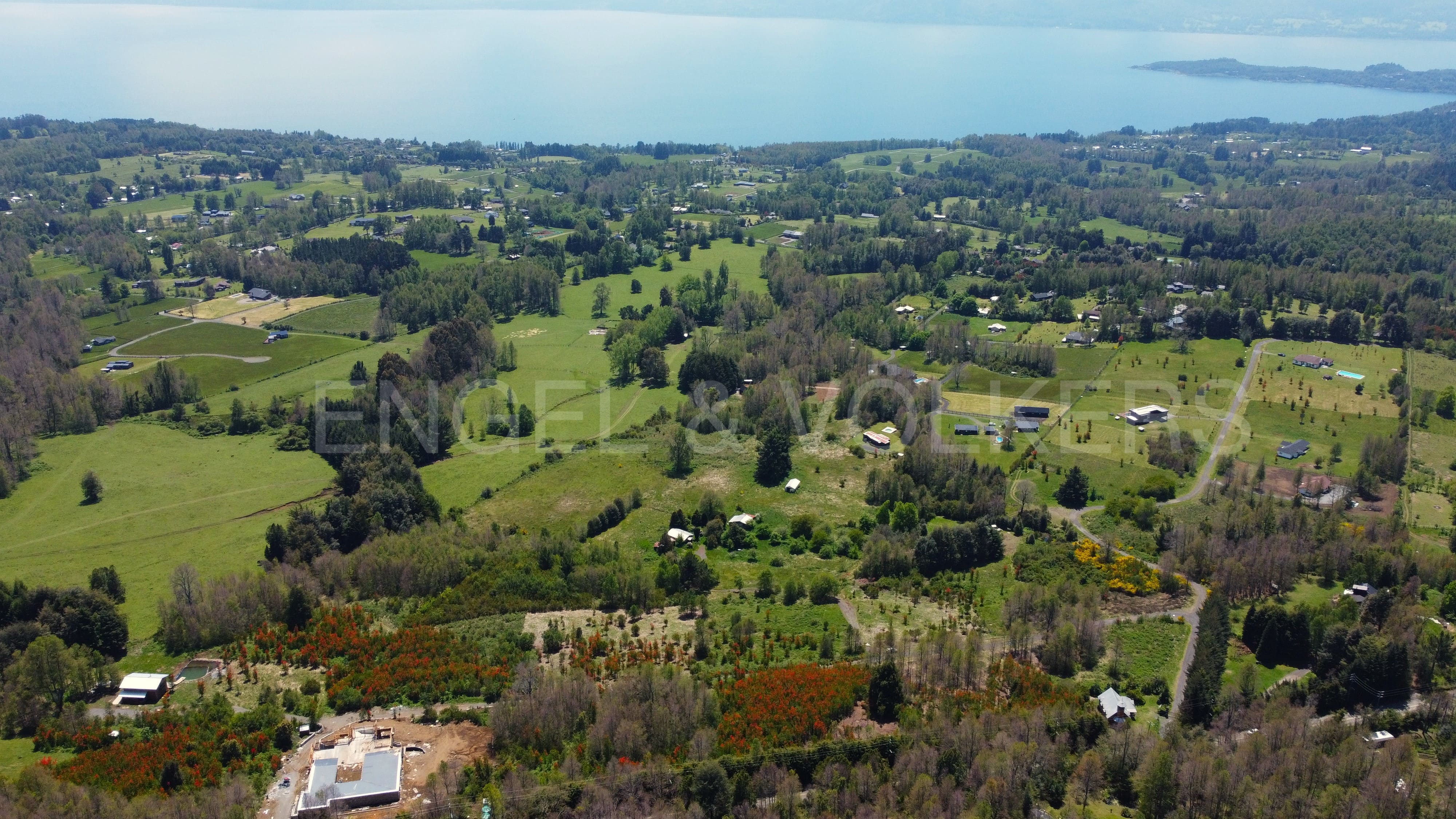 Aerial view of a lush green landscape with scattered houses, trees, and a large lake in the distance under a bright sky. [AI-generated text]