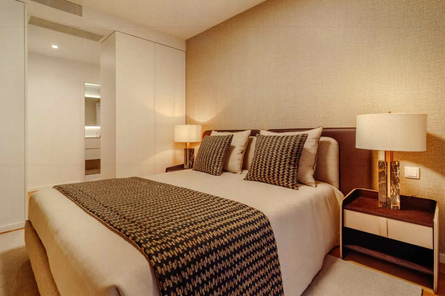 Bedroom with a queen bed, patterned pillows and throw, nightstand with lamp, and a glimpse of a bathroom through an open doorway.