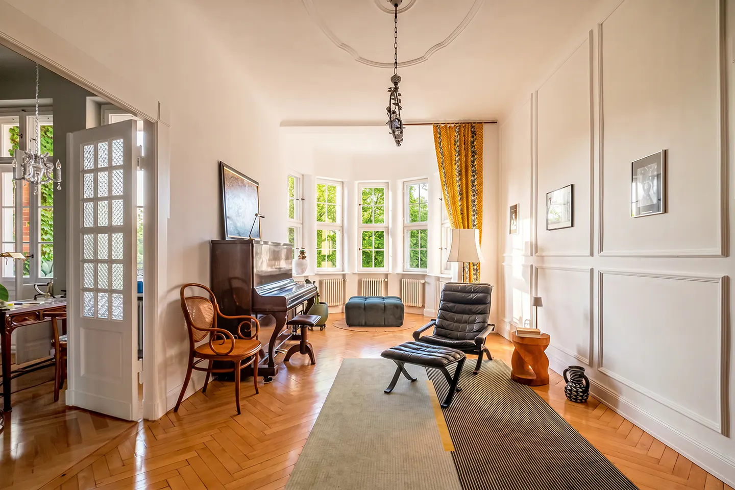Bright living room with herringbone wood floors, piano, black leather chair, and bay window with yellow curtains.