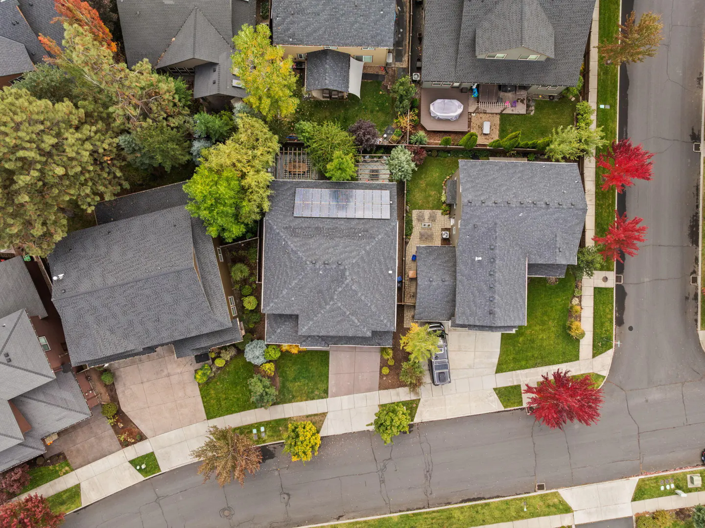 Aerial view of a residential neighborhood with gray roofs, green lawns, and colorful trees in autumn. Solar panels are visible on one roof.