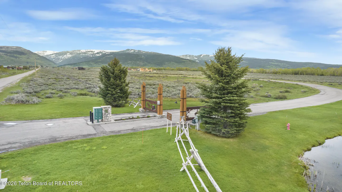 Gated entrance to a property with green grass, trees, and mountains in the background under a blue sky with white clouds.