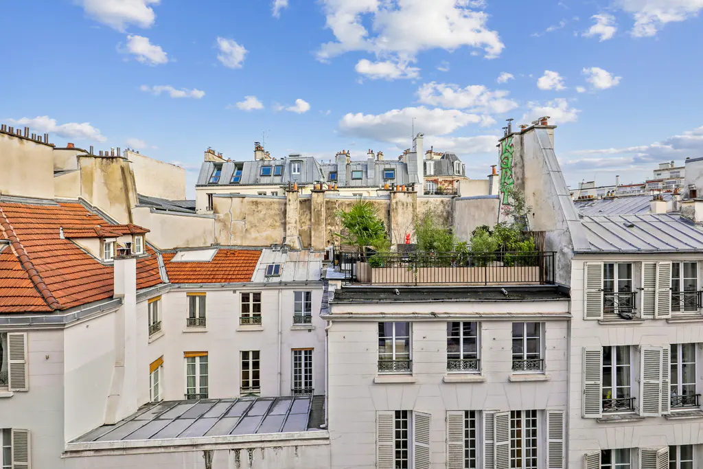 Parisian rooftops with white buildings, red tile roofs, and a rooftop garden under a blue sky with clouds.