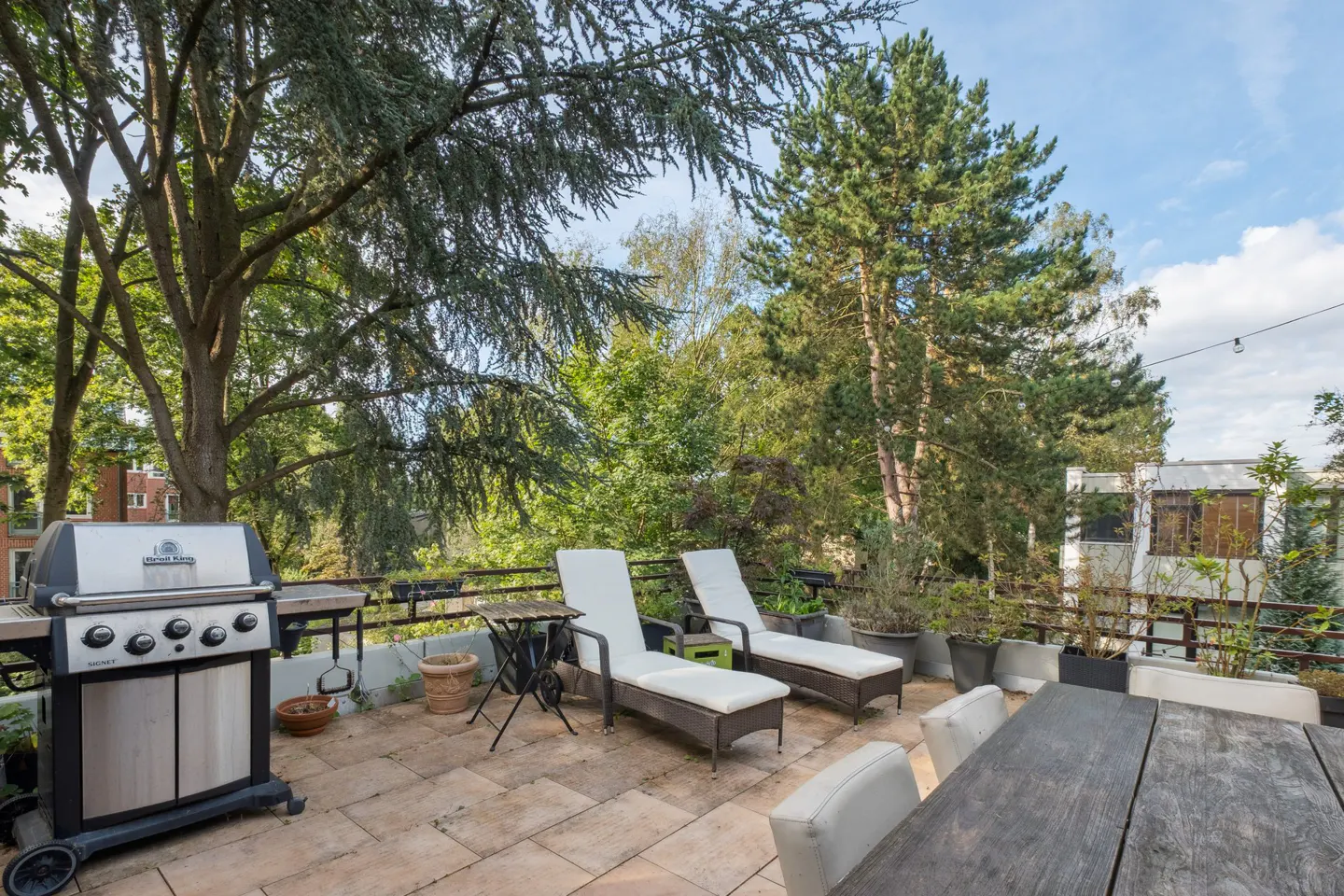 Outdoor patio with grill, lounge chairs, and dining table. Lush green trees surround the space.