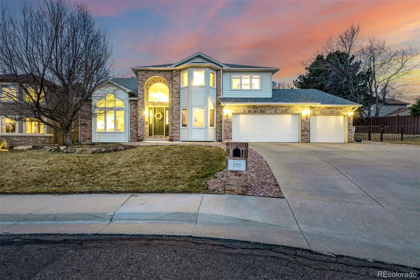 Two-story house with white siding, brick accents, and a two-car garage at dusk. A green wreath hangs on the front door.