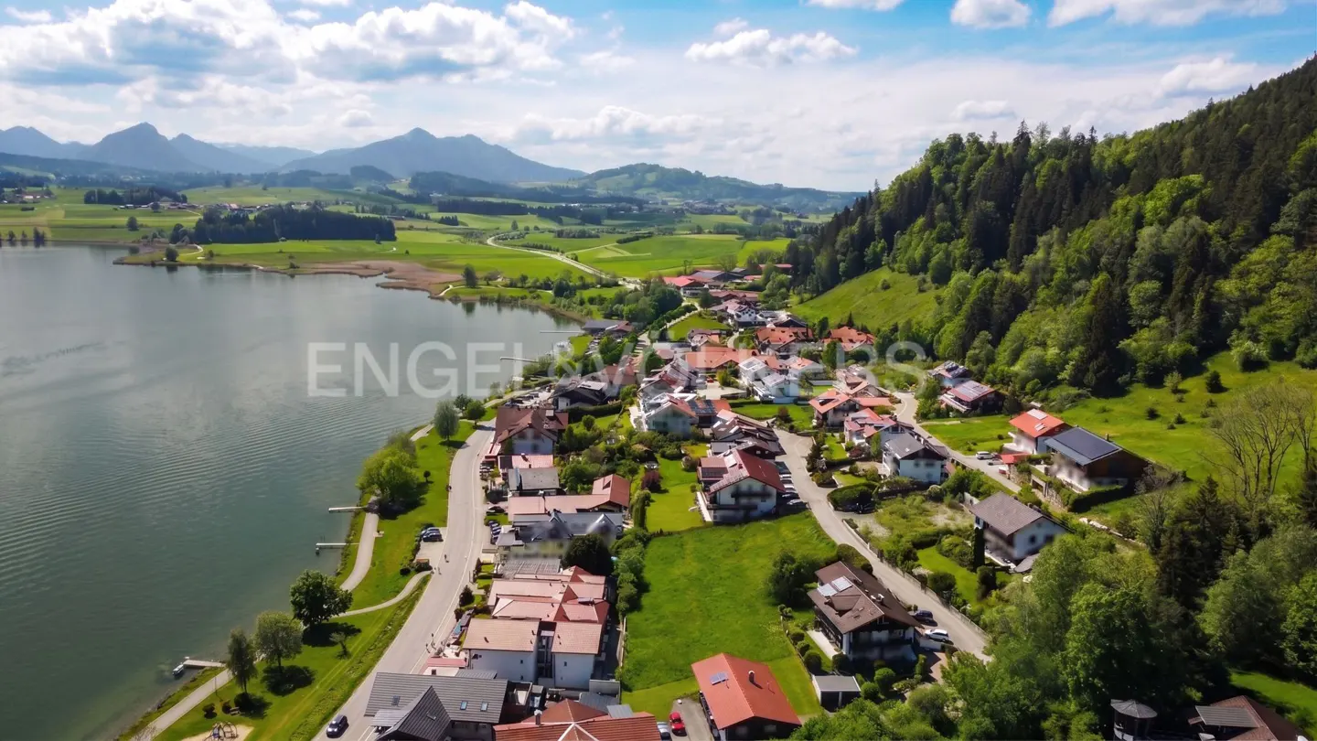 Aerial view of a lakeside village with red-roofed houses, green hills, and mountains under a blue sky.