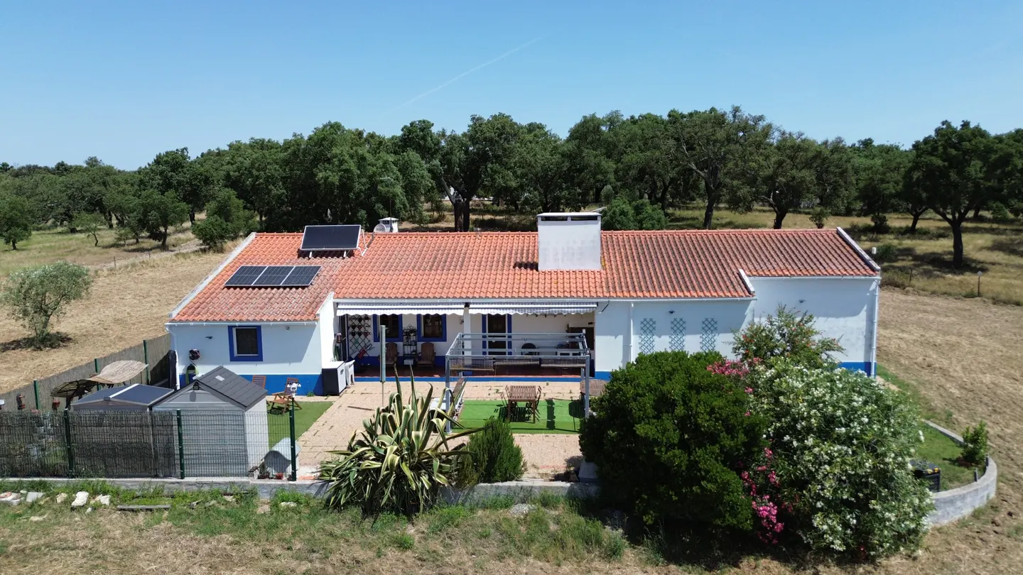 Aerial view of a white single-story house with a red tile roof, solar panels, and a patio in a rural, tree-lined landscape.