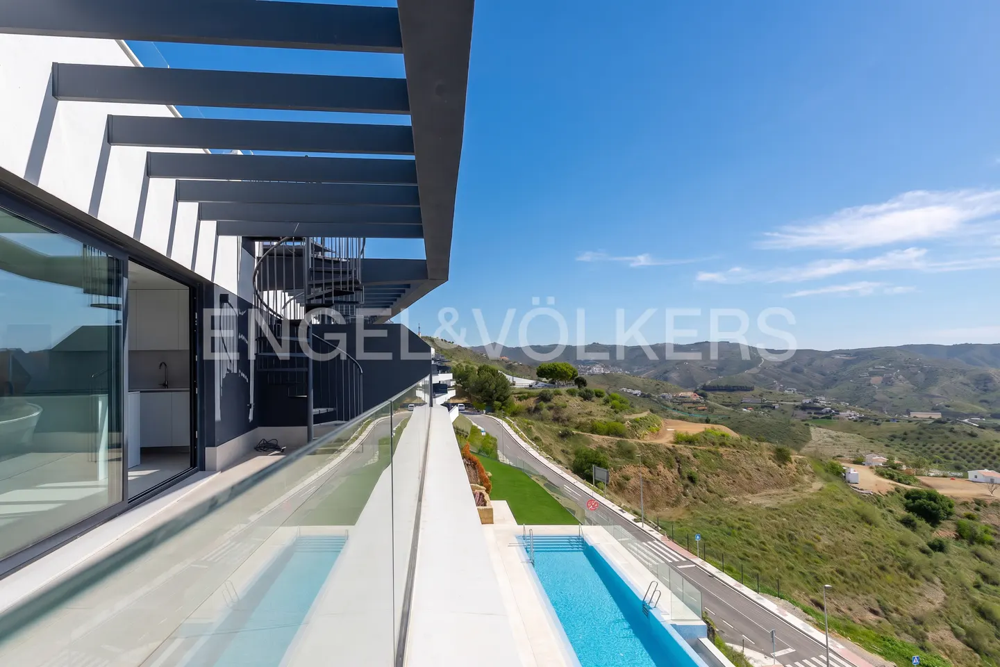 Balcony view of a modern home with a pool. The home has a spiral staircase and a gray pergola. Rolling hills are in the background.