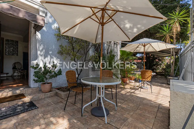 Outdoor patio with a round table, wicker chairs, and a large white umbrella. Plants and another umbrella are in the background.