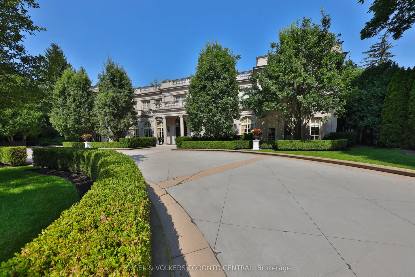 Beige mansion with white columns and a circular driveway lined with green hedges and trees under a blue sky.