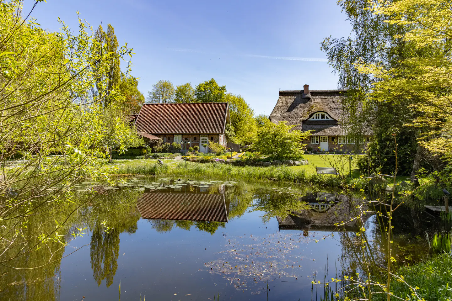 Two traditional houses with tiled and thatched roofs sit behind a calm pond reflecting the buildings and blue sky.