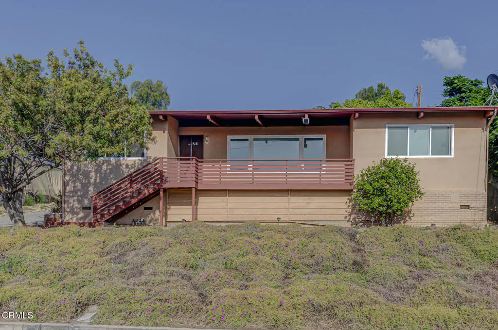 Tan two-story house with a brown roof, deck, and stairs. The house sits on a hill covered in green and purple ground cover.