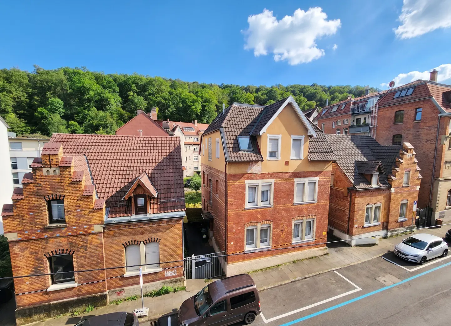 View of red brick buildings with pitched roofs, parked cars, and green trees under a blue sky.