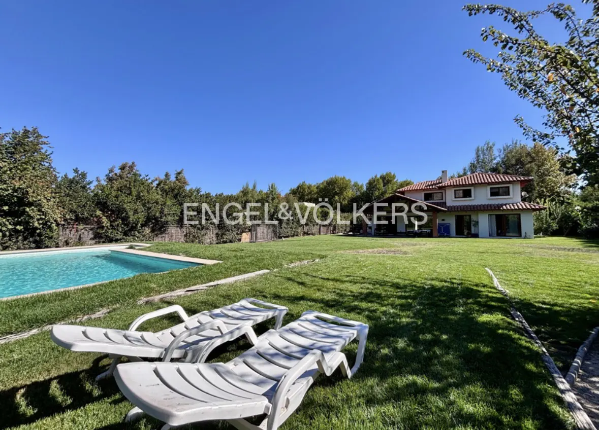 Two white lounge chairs sit on a green lawn, with a pool and house in the background under a blue sky.