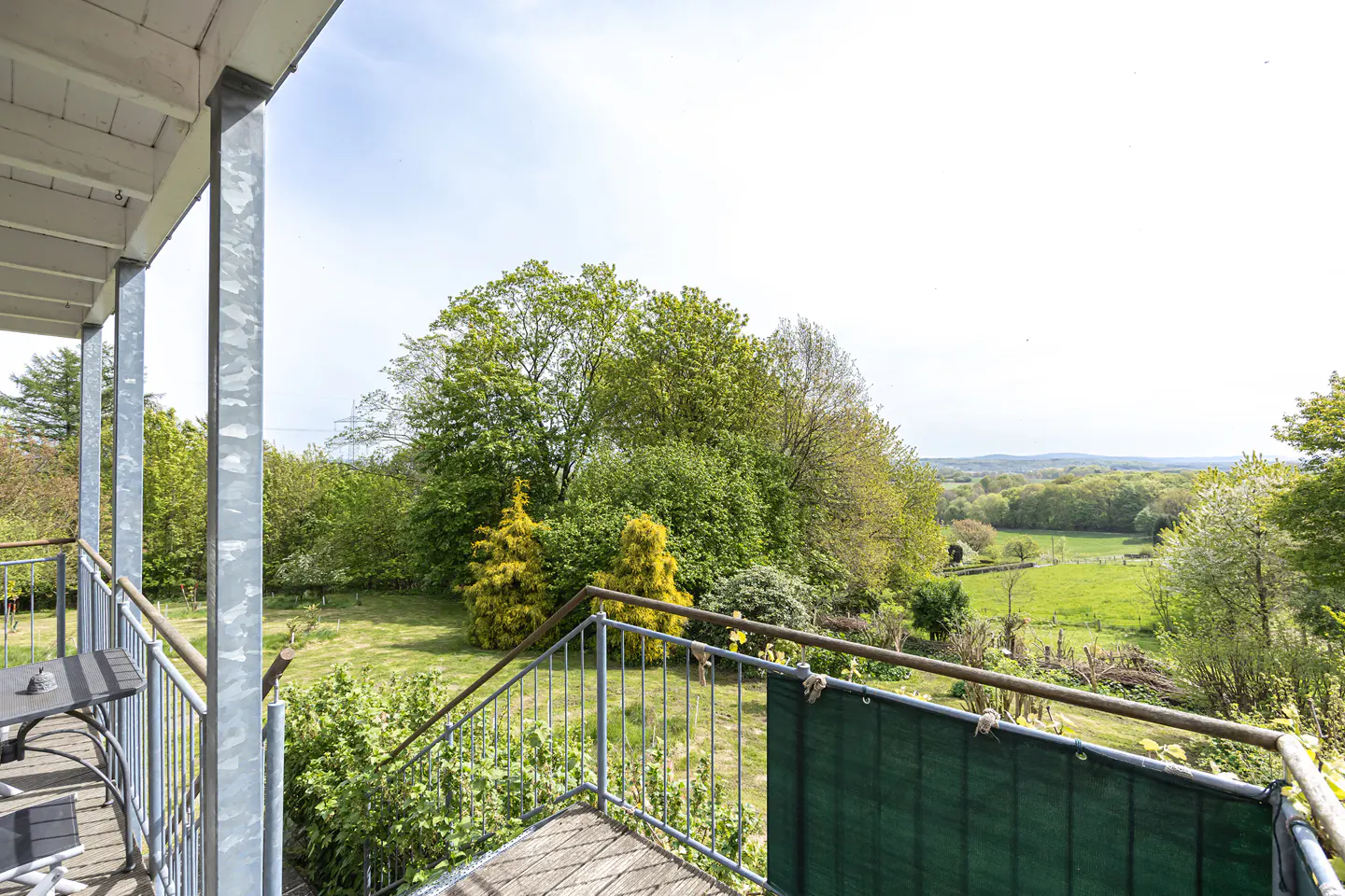 View from a balcony with metal railings overlooking a green lawn and trees on a sunny day.