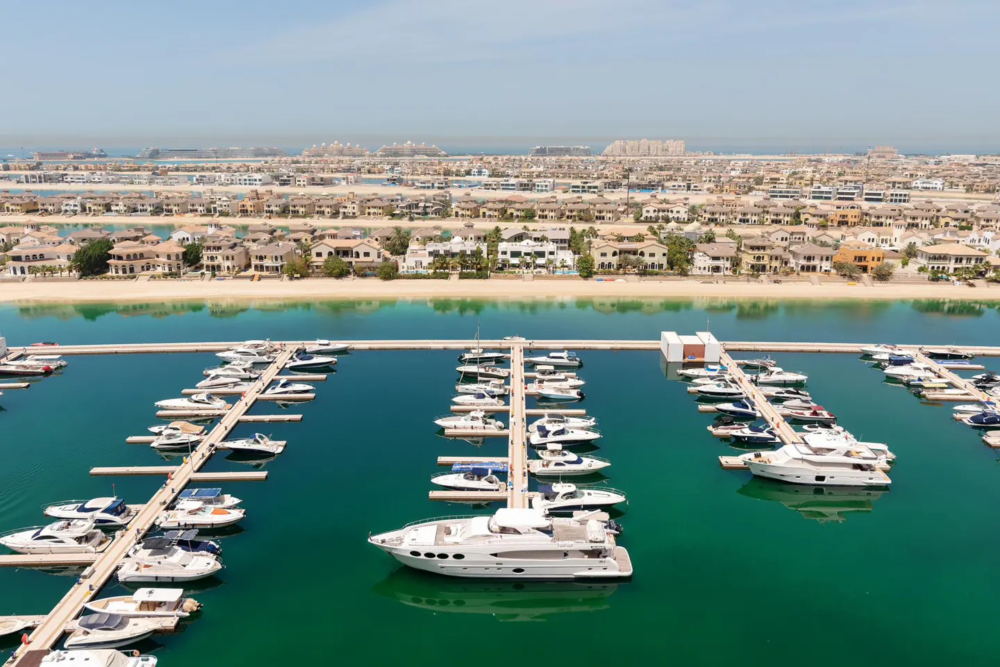 Aerial view of a marina filled with white yachts, turquoise water, and luxury homes on a sandy shore under a clear blue sky.