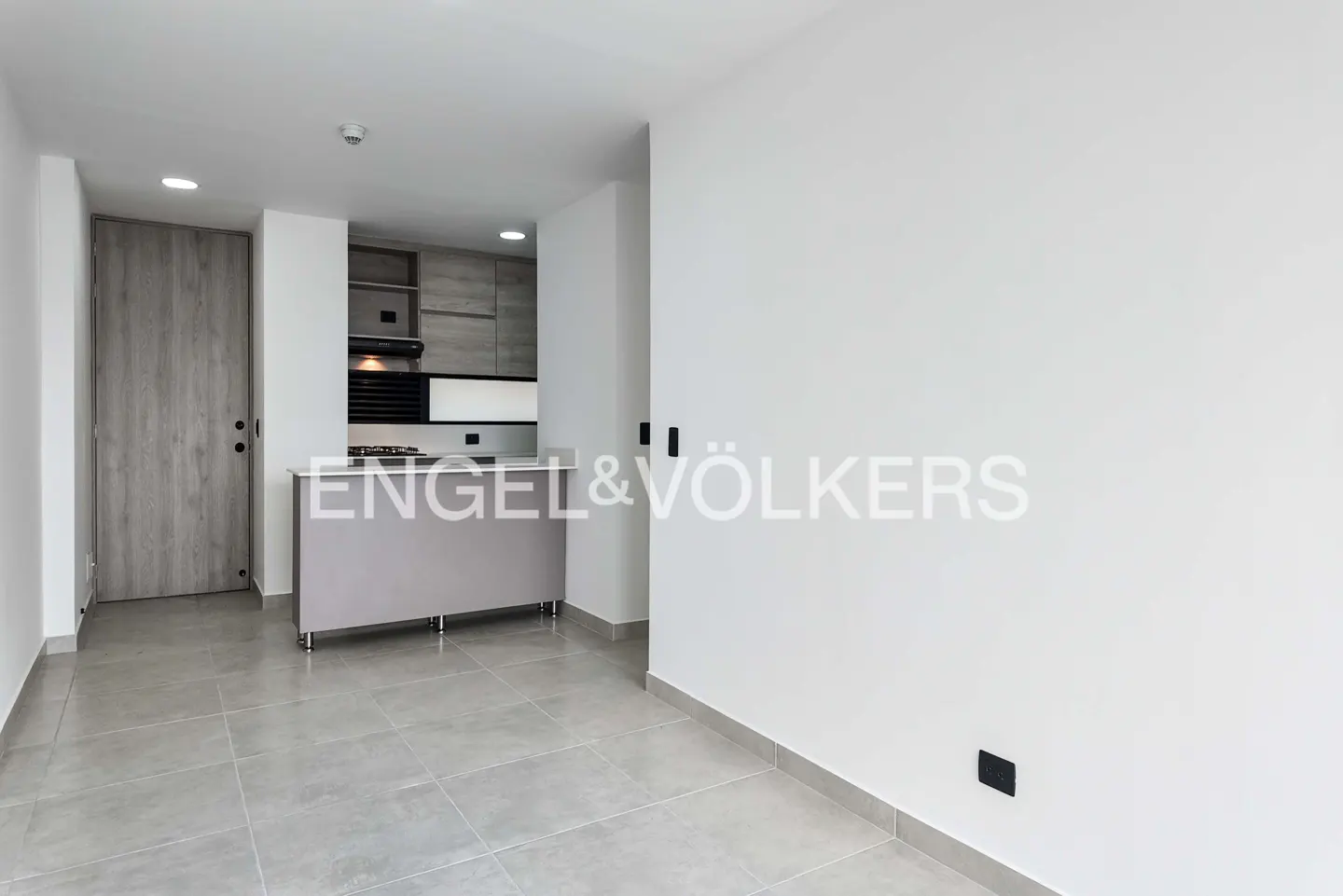A bright, empty apartment with gray tile floors, white walls, and a small kitchen area. A gray door is on the left.