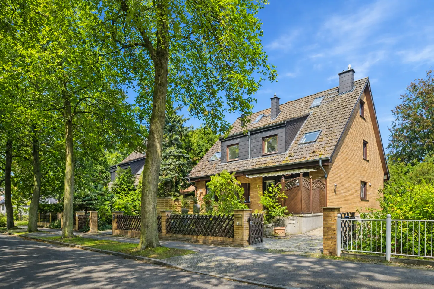 Two-story brick house with a gray roof and dormer windows, surrounded by green trees and a fence.