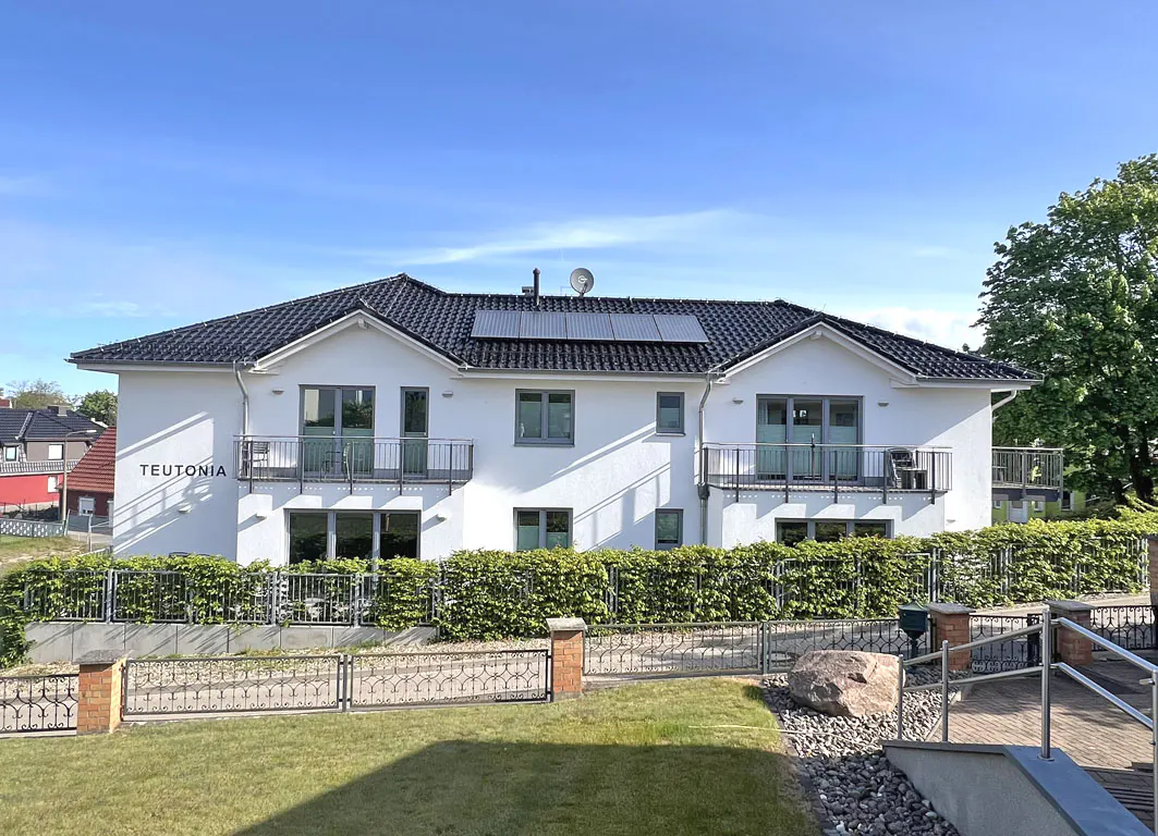 Two-story white building with black roof and solar panels. Balconies, green hedge, and lawn. "Teutonia" sign on the left side.