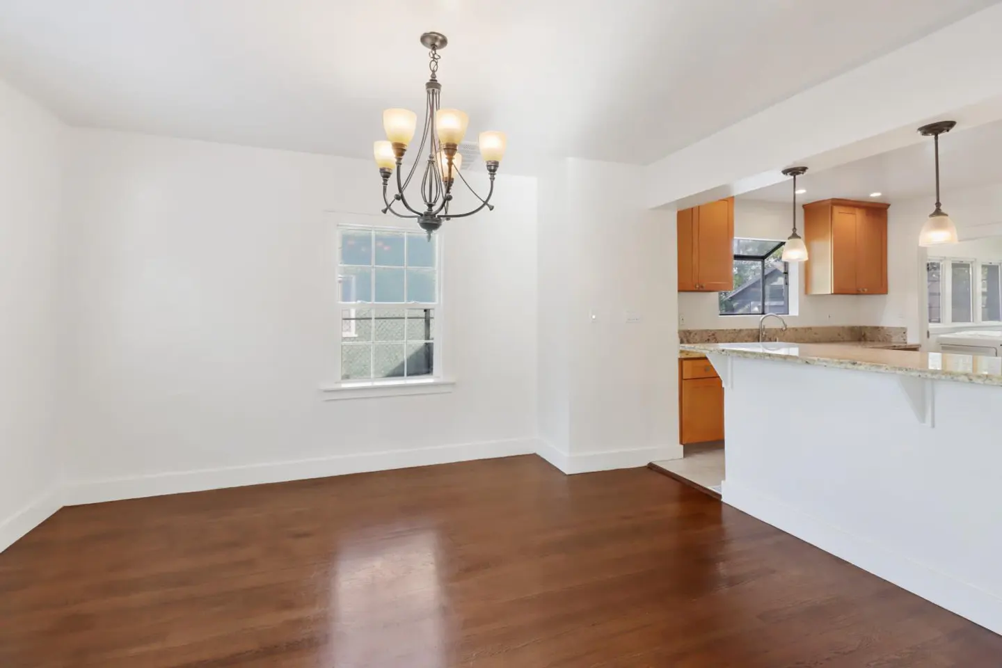 Bright, empty dining room with hardwood floors, white walls, and a chandelier. Kitchen visible through opening with wood cabinets and granite countertops.