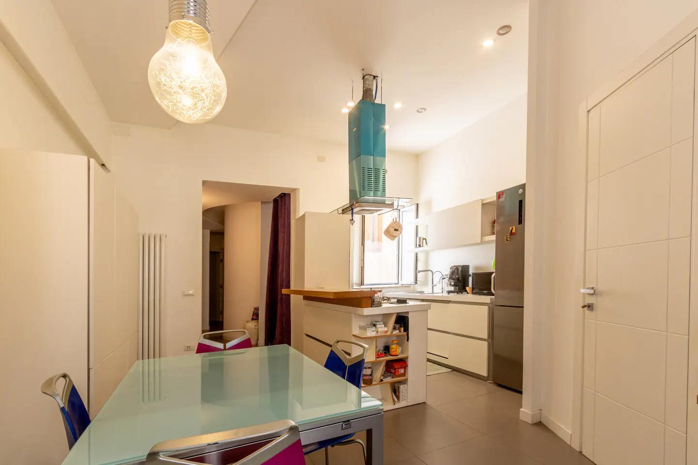 Modern kitchen with a glass dining table, blue chairs, a teal range hood, and stainless steel refrigerator.